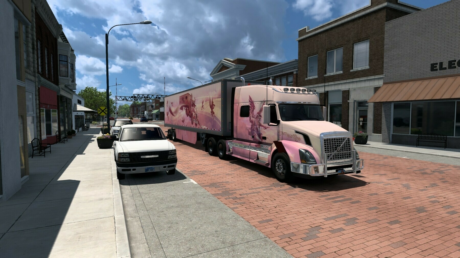 A pink semi-truck with designs on its trailer is parked on a brick-paved street alongside parked cars and buildings.