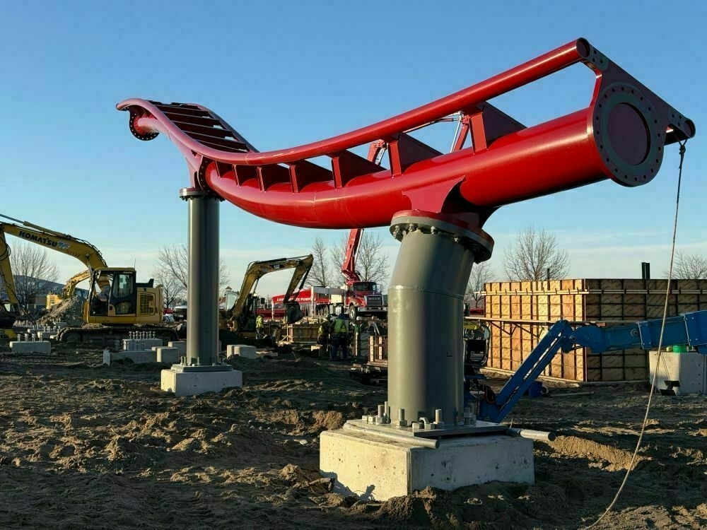 The image shows a construction site with a partially installed red roller coaster track section. The track is supported by large gray steel columns anchored to concrete footings. One of the columns appears to be tilted, possibly as part of the track's design. Various construction equipment, including excavators and cranes, are present, along with workers wearing safety gear. The background features trees and a clear blue sky, suggesting a sunny day. The construction site appears to be in the early stages, with foundational work still underway.