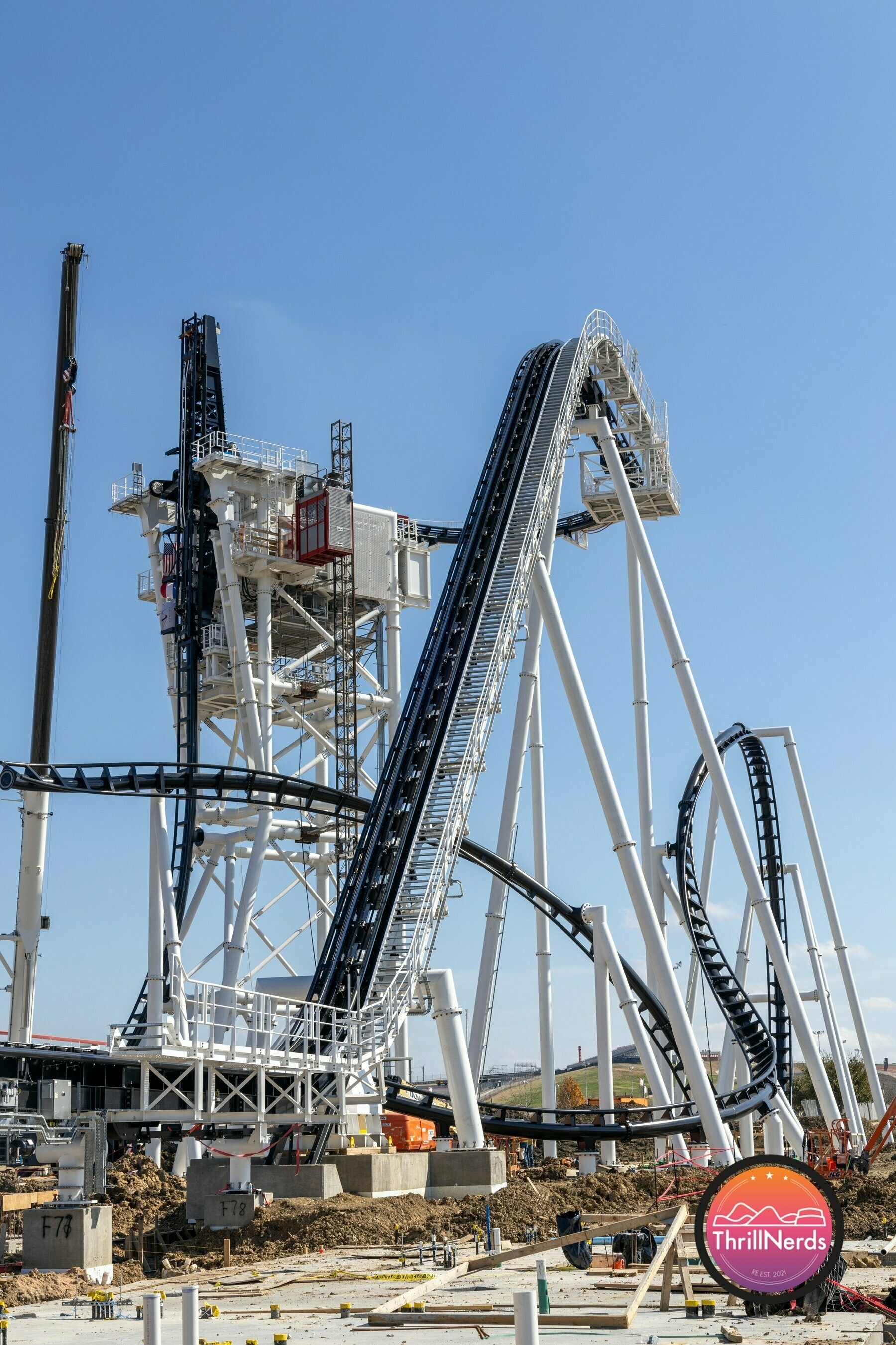 A large roller coaster under construction features white and black tracks with a steep incline and metallic support structures.