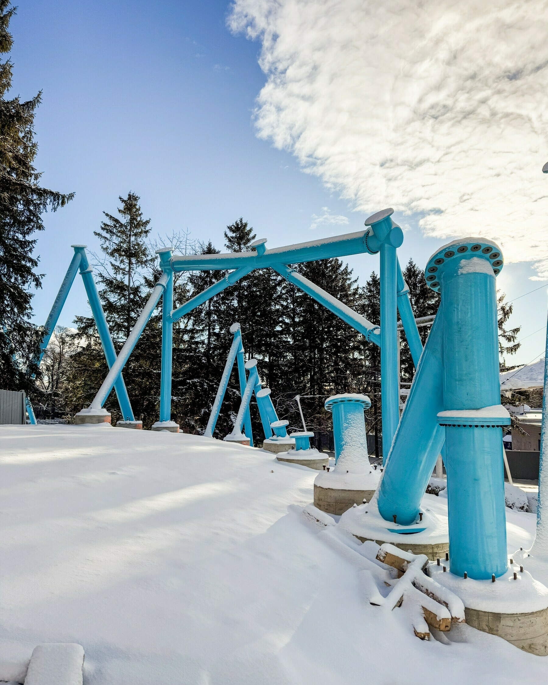 Bright blue metal supports stand amidst a snowy landscape with tall evergreen trees and a partly cloudy sky.