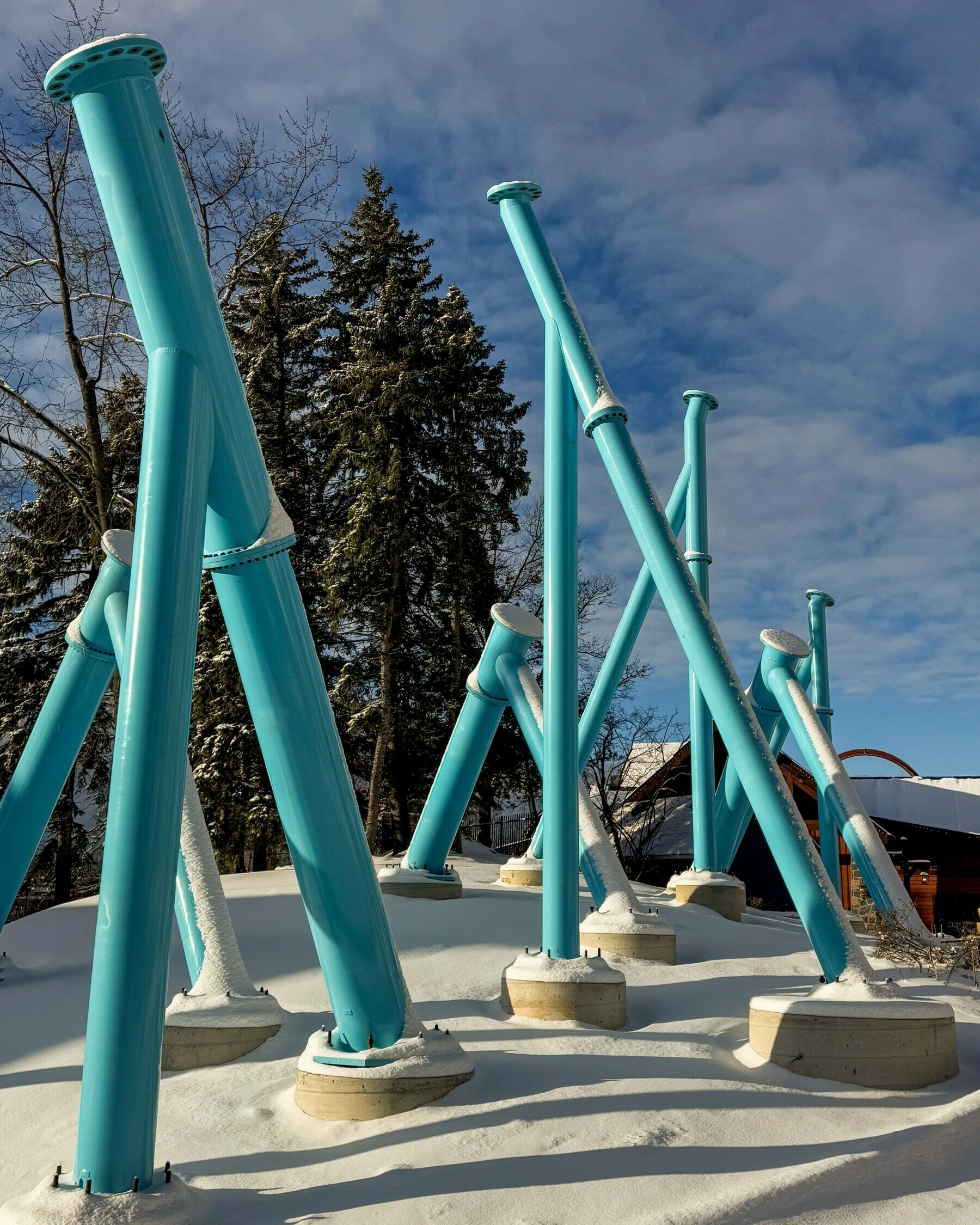 Several large, turquoise steel beams are anchored in a snow-covered landscape, positioned at various angles with trees and a building in the background.