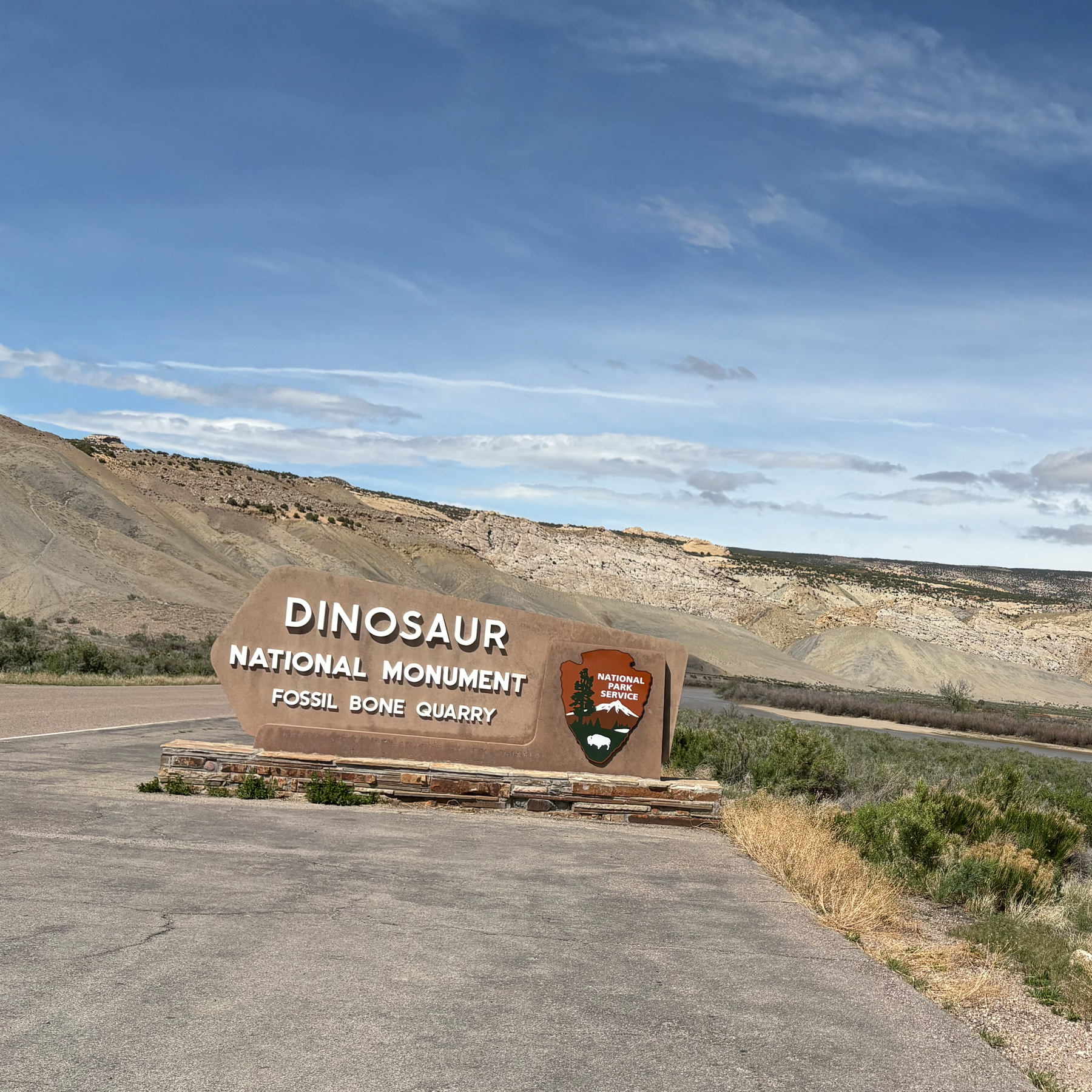 A sign for Dinosaur National Monument's Fossil Bone Quarry is set against a backdrop of arid, rocky hills under a partly cloudy sky.