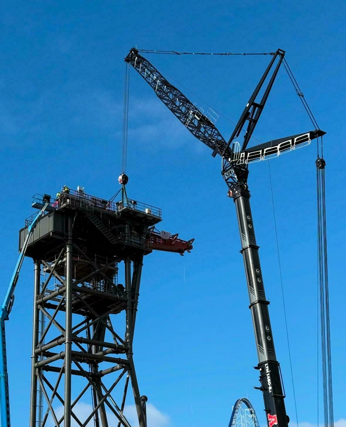 A large crane is lifting a segment of a roller coaster against a clear blue sky.