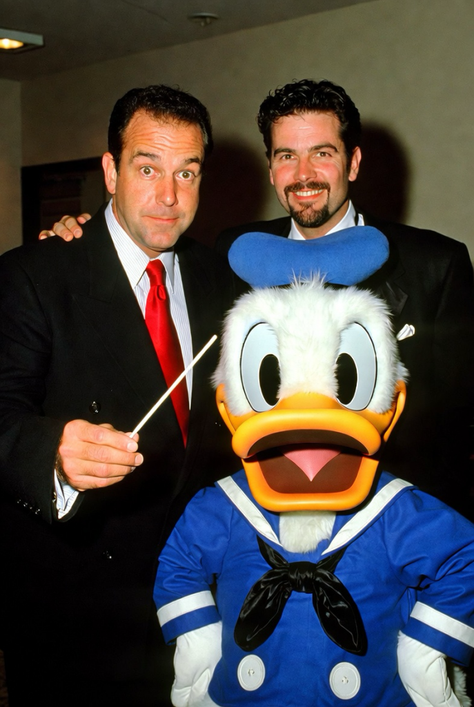 Two men in suits stand indoors beside a person dressed in a full Donald Duck costume. One man smiles while holding a conductor’s baton, and the other stands just behind the character, also smiling. Donald Duck wears his classic blue sailor outfit, black bow tie, and blue hat. The scene appears to be at an event or gathering, with all three posing for the photo.