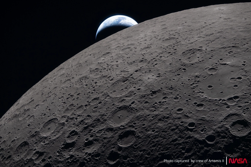 A high‑resolution photograph shows the Moon’s cratered surface in the foreground, lit sharply by sunlight that highlights its rough texture. Above the lunar horizon, Earth appears as a bright blue‑and‑white sphere rising against the blackness of space. Text on the image reads “Photo captured by crew of Artemis II | NASA.”